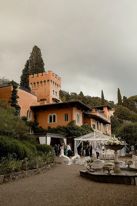 Outdoor wedding reception under a clear tent wedding reception with string lights, white chairs, and a stone fountain in a villa courtyard