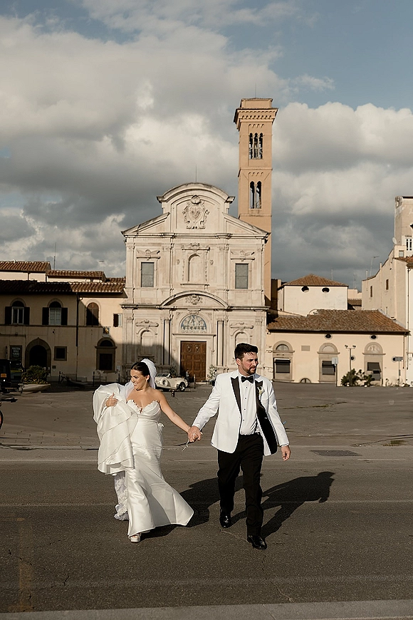 Couple portrait of newlyweds holding hands, bride lifting her dress and veil beside a church facade and bell tower in a town square