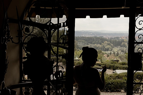 Getting ready moment as a bride in her wedding dress eats a snack by an open doorway, looking out to garden shrubs and distant hills