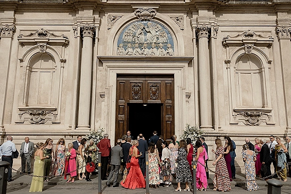 Wedding guests outside church in suits and long dresses mingle on stone steps by ornate doors, one holding a bouquet near floral aisle arrangements
