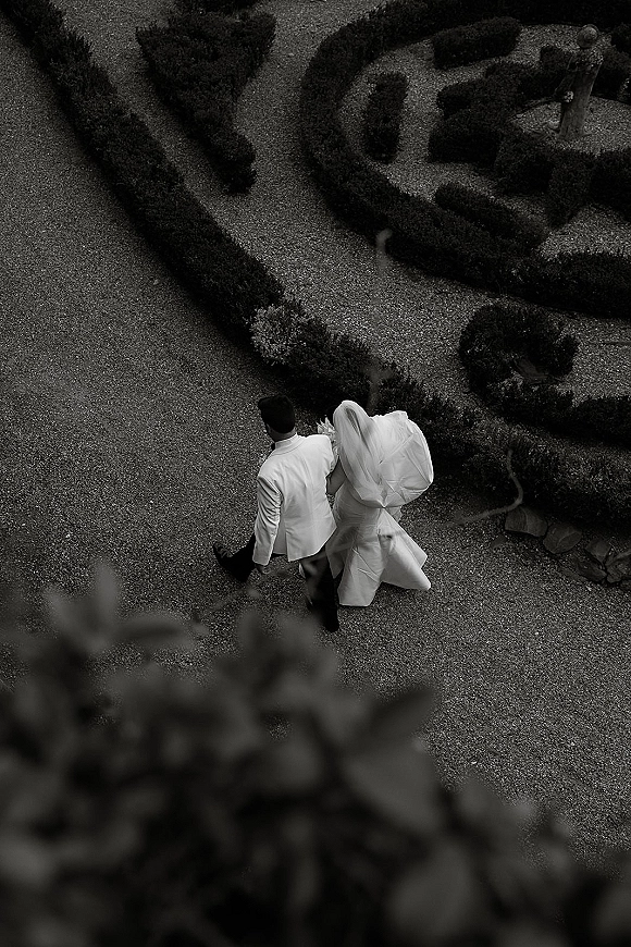 Newlywed portrait from above of bride and groom walking away, veil trailing, holding a bouquet on a gravel path in a garden maze