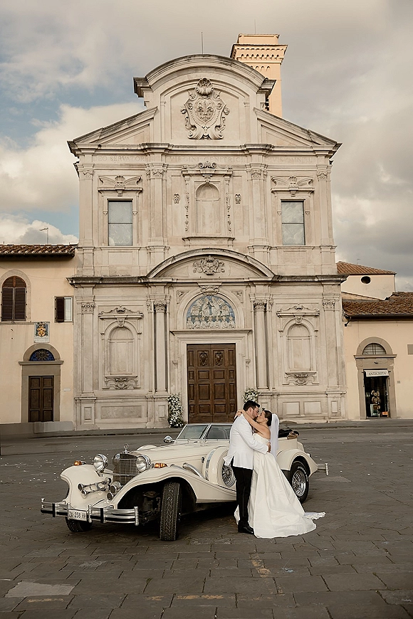 Wedding couple portrait of bride and groom kissing beside a vintage convertible car, veil trailing on stone plaza outside a church facade