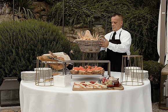 Wedding cocktail hour buffet table with charcuterie board, bread and fruit platters on white linen, set on an outdoor patio by hedges