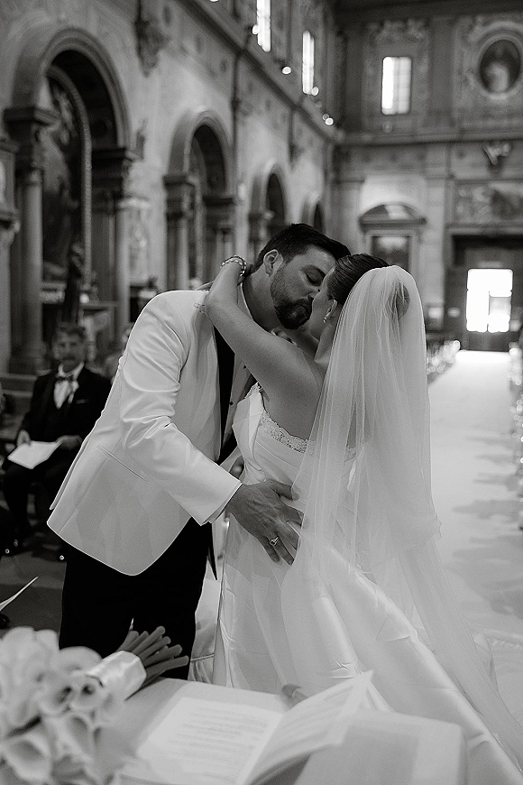 Wedding kiss as bride in strapless lace gown and long veil embraces groom in white tuxedo jacket along church aisle runner near altar