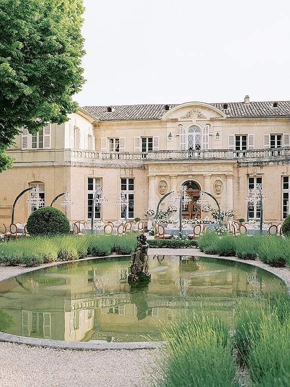 Outdoor reception setup with round tables and white tablecloths beneath crystal chandeliers in a stone manor courtyard by a reflecting pool fountain