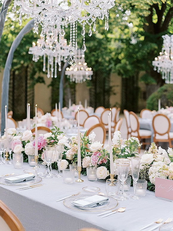 Reception tablescape with an outdoor reception table set in a garden courtyard, lined with white taper candles, blush florals, gold flatware and chandeliers overhead