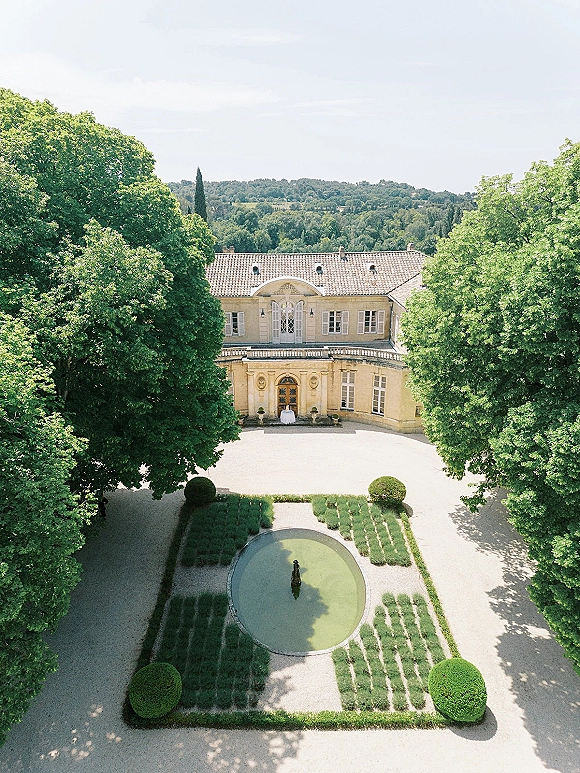 Wedding venue exterior of a stone manor house with arched doorway, balcony, and circular fountain in a formal gravel courtyard