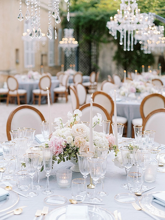 Reception tablescape for an outdoor wedding reception with blush florals, taper candles, crystal chandeliers, and gold flatware in a courtyard