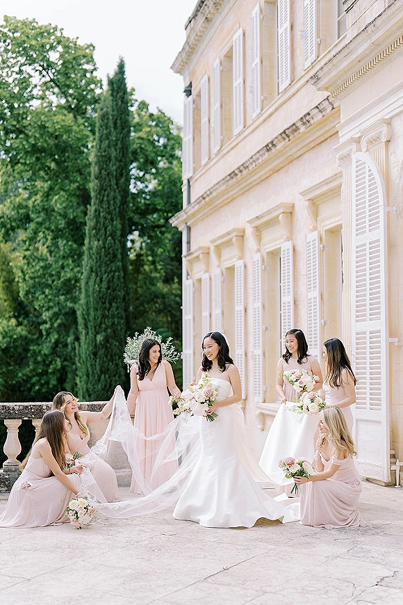 Bride with bridesmaids in a bridal party portrait, holding bouquets and arranging her long veil on a stone terrace by an estate facade