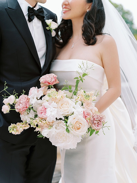 Couple portrait of bride and groom, bride holding bouquet with long veil beside groom in black tuxedo, outdoor greenery and sky
