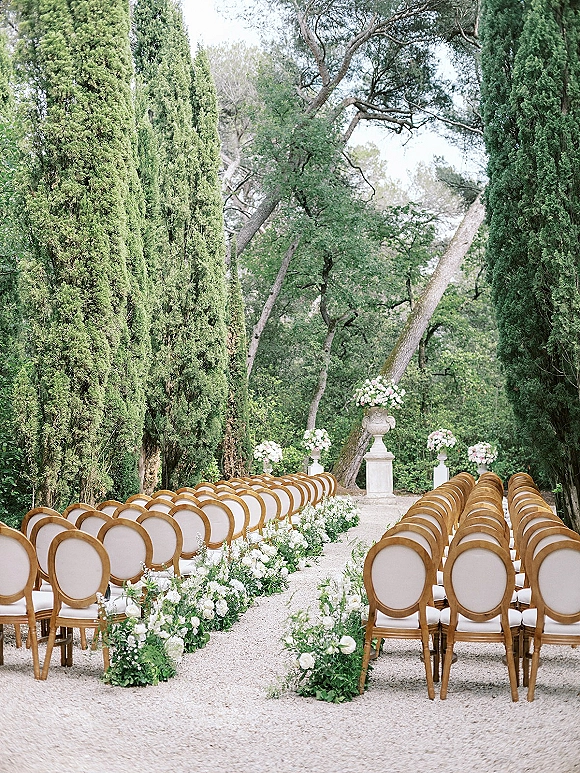 Ceremony aisle design with outdoor garden ceremony seating, white rose and greenery aisle flowers lining a gravel path with stone urns