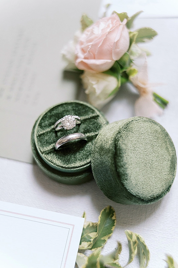 Wedding rings with a diamond engagement ring close up resting on a green velvet ring box beside a blush rose, greenery, and invitation on a light tabletop