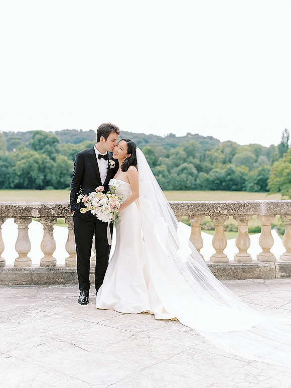 Couple portrait of groom kissing bride’s forehead as she holds a bouquet, her long veil flowing on a stone terrace with balustrade and trees