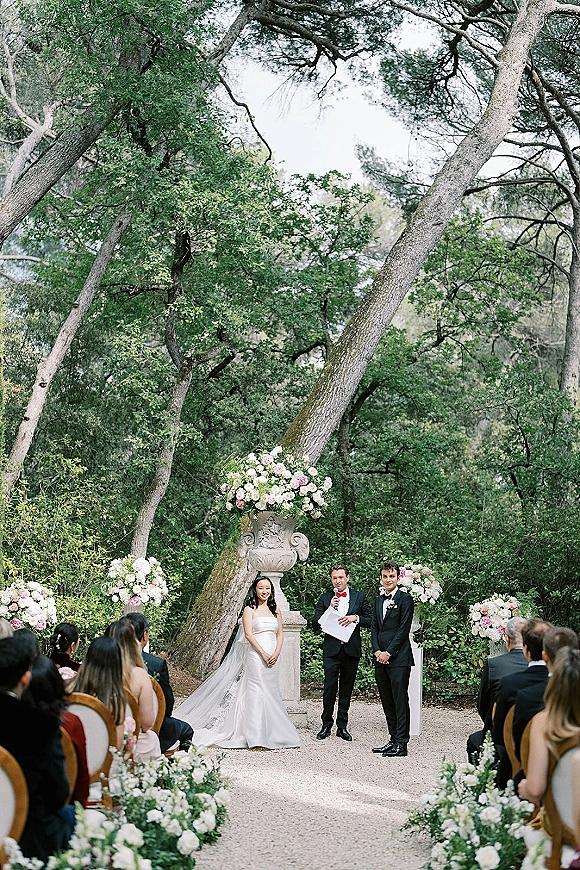 Ceremony moment as bride and groom stand with officiant at a garden altar, framed by white and blush floral urns under tall trees