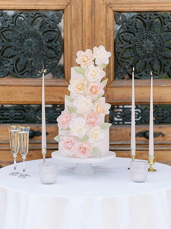 Wedding cake with sugar flower details on a stand, flanked by taper candles and champagne flutes on a white cloth before ornate doors