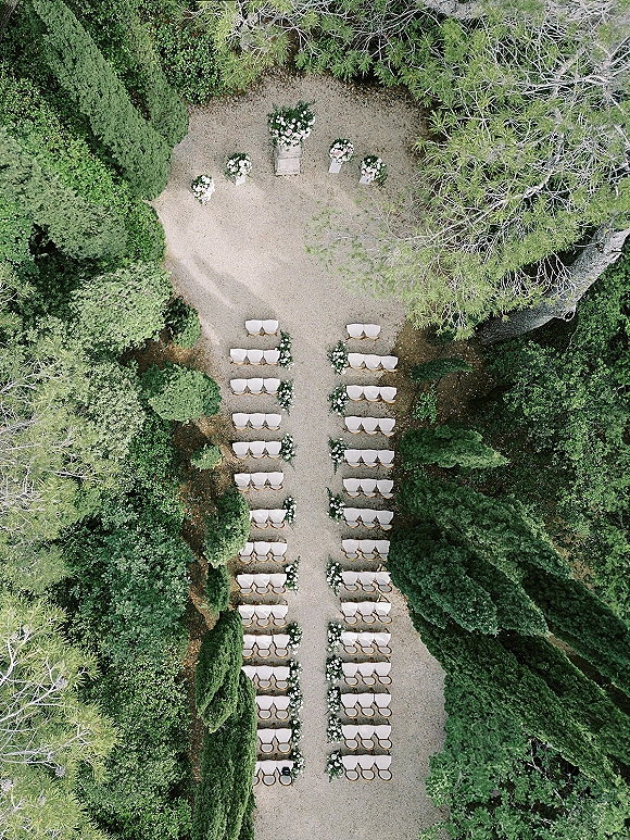 Ceremony setup for an outdoor wedding ceremony with white chairs in rows, a gravel aisle, and floral markers in a garden clearing