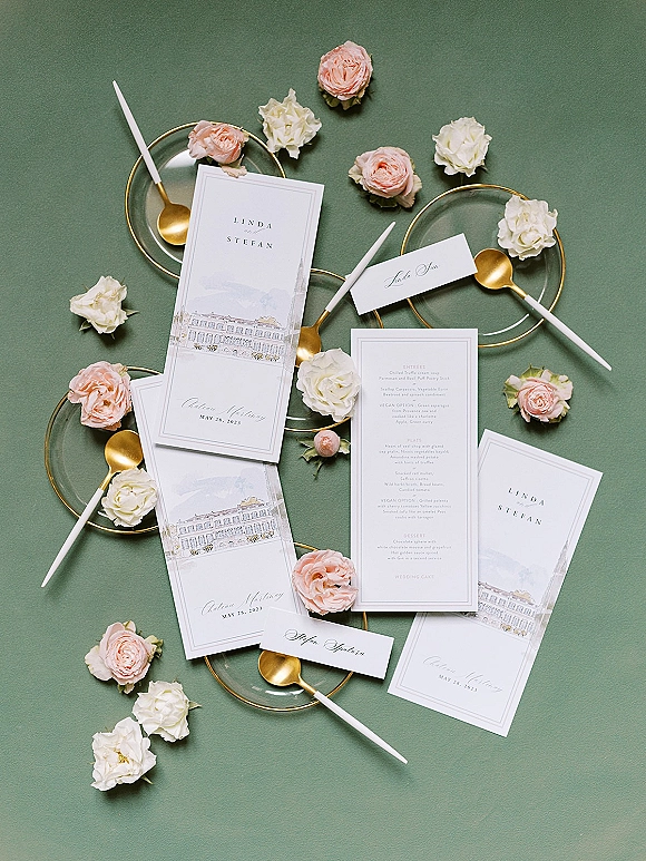 Invitation suite wedding stationery flat lay with RSVP and menu cards, gold-rim plates and spoons, taper candles, blush roses on green backdrop