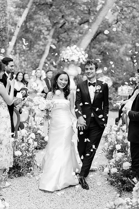 Wedding recessional as newlyweds walk hand in hand down a gravel garden aisle, guests tossing petals, bride with bouquet, groom in tuxedo