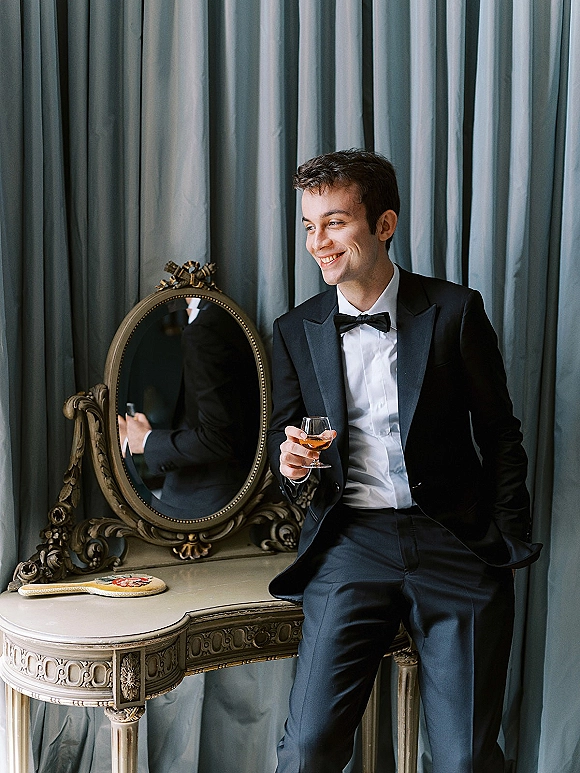 Groom portrait in tuxedo holding a drink, smiling by an ornate mirror and console table against blue drapery curtains