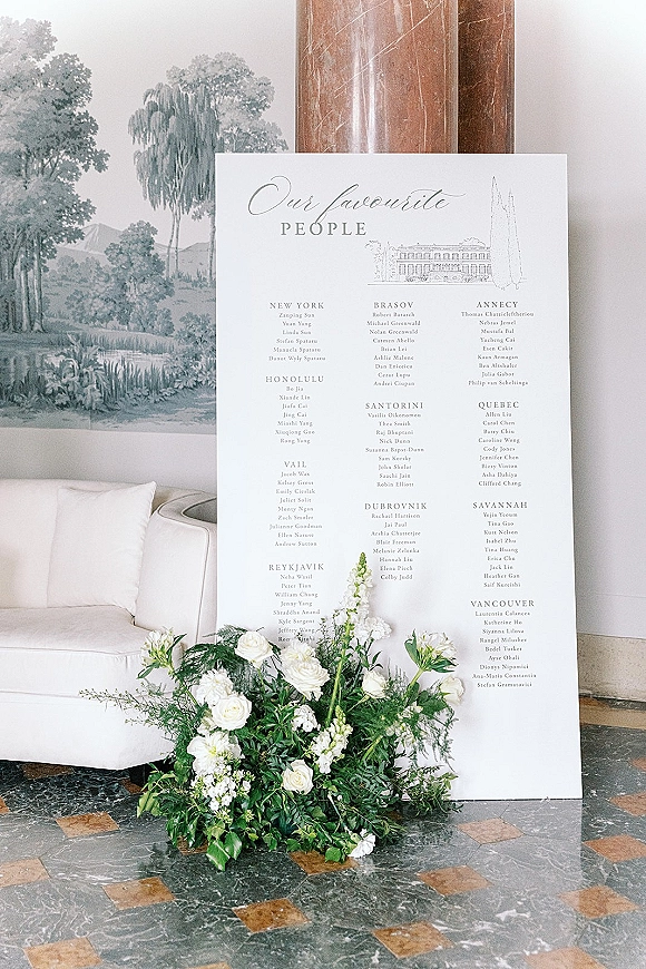 Wedding seating chart with calligraphy seating chart lettering on a large board, styled with white roses and greenery by a marble column
