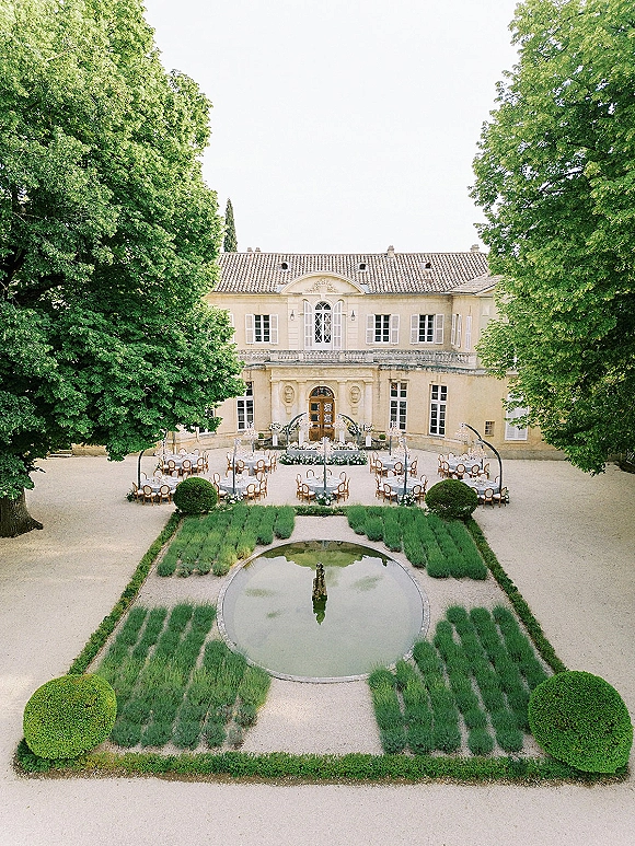 Outdoor reception setup with round tables in white linens and bistro chairs, floral centerpieces around a courtyard fountain by a chateau facade