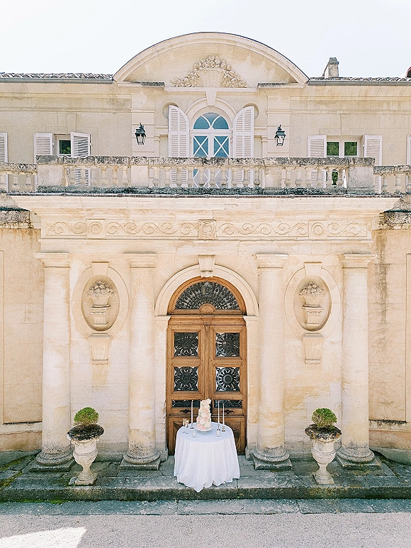 Wedding cake table with a tiered cake and taper candles on a round white tablecloth, set before a stone villa with wooden doors