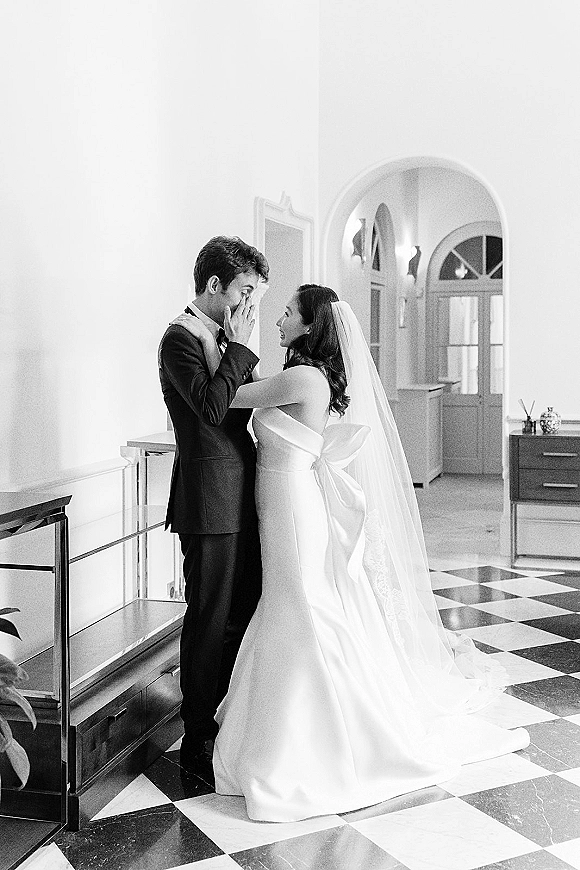First look moment as the bride in a strapless bow-back gown and veil cups the groom’s face, his tearful reaction in an arched hallway