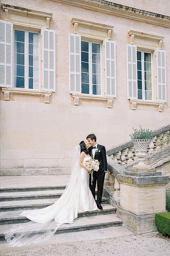 Wedding kiss portrait of bride and groom kissing on stone steps, her veil and long train flowing, holding a pastel bouquet by balustrade