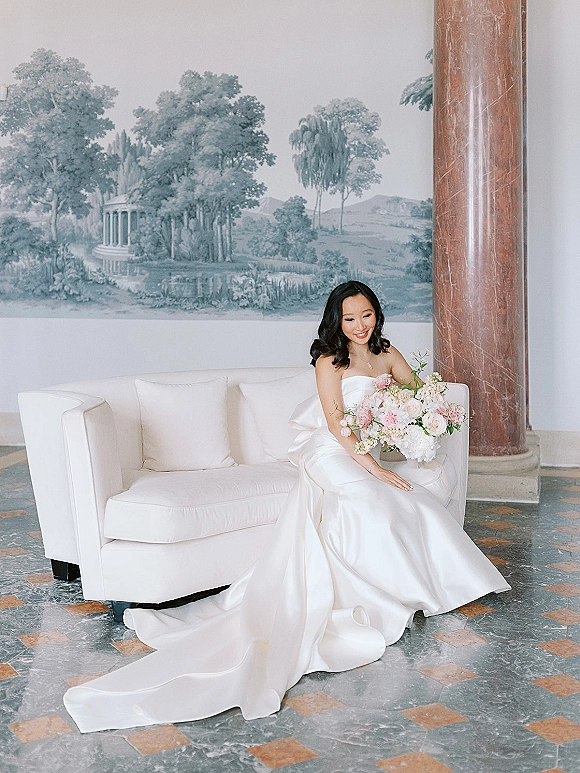 Bridal portrait of a bride sitting on a sofa in a strapless satin wedding dress, holding a pink and white bouquet in an indoor lobby