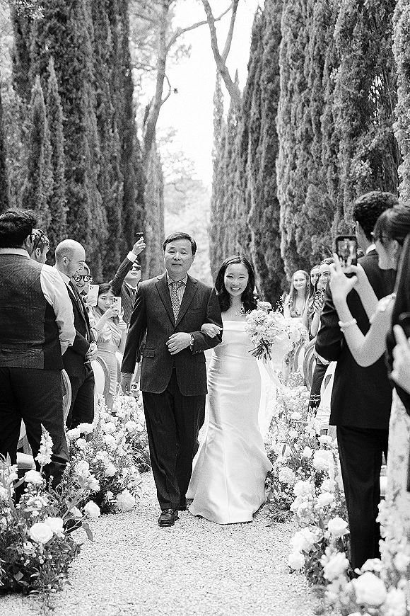 Bridal processional with bride walking down aisle escorted by father, holding white bouquet along flower-lined gravel path under cypress trees