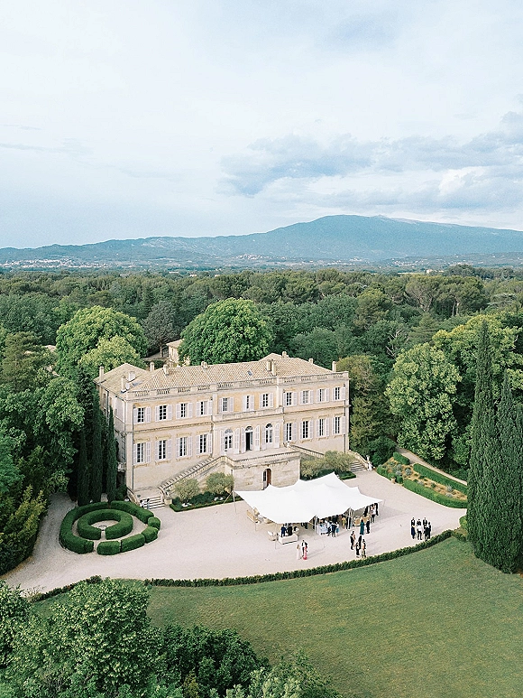 Wedding reception tent set in a gravel courtyard with cocktail tables beside a chateau and manicured hedges, mountains beyond