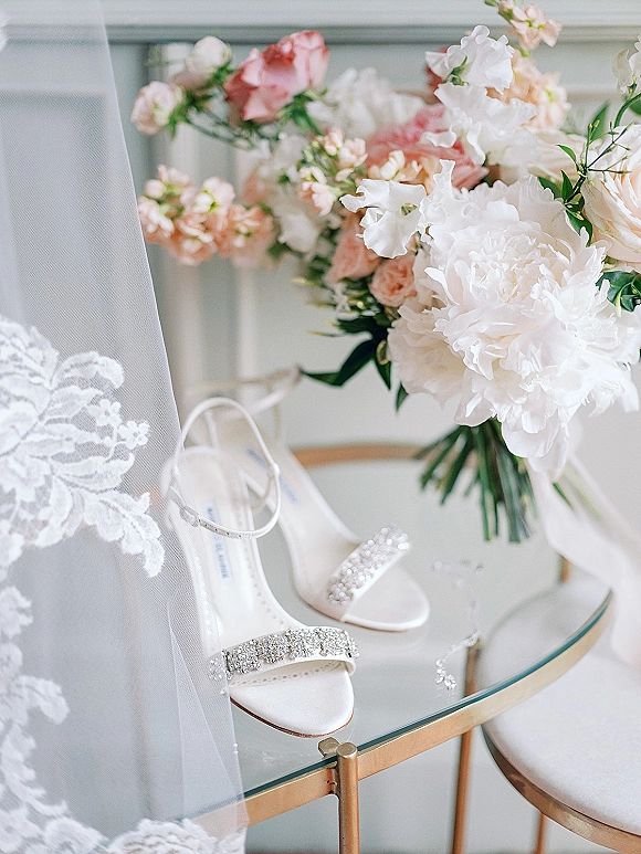 Bridal accessories flat lay with rhinestone bridal heels beside lace veil and peony-rose bouquet on a gold-framed glass table