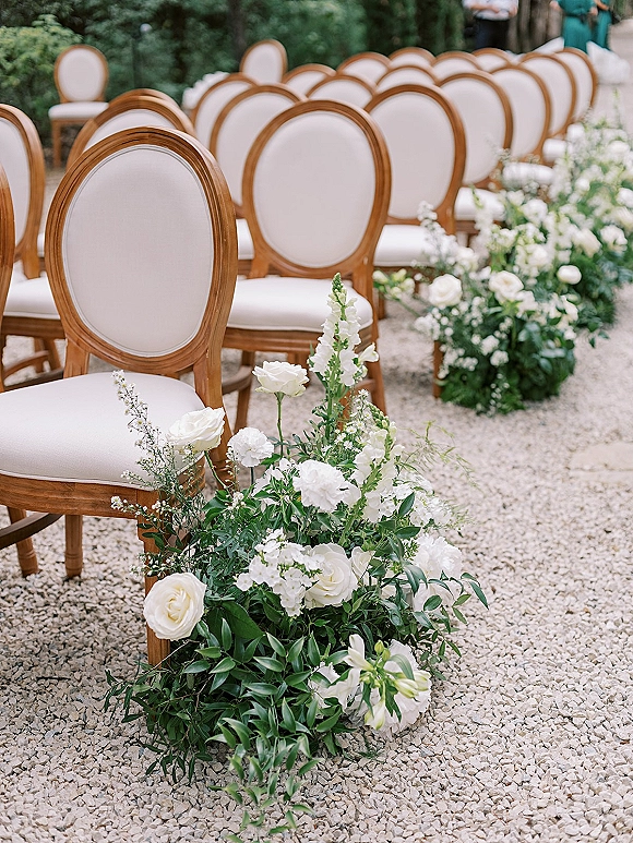Ceremony aisle decor with white rose and greenery florals lining a gravel walkway, flanked by wood frame and white upholstered chairs in garden hedges