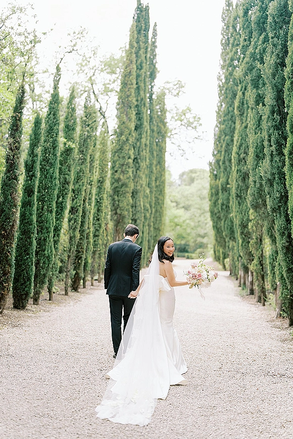 Couple portrait of bride and groom walking away hand in hand, bride looking back with bouquet on a cypress-lined gravel path