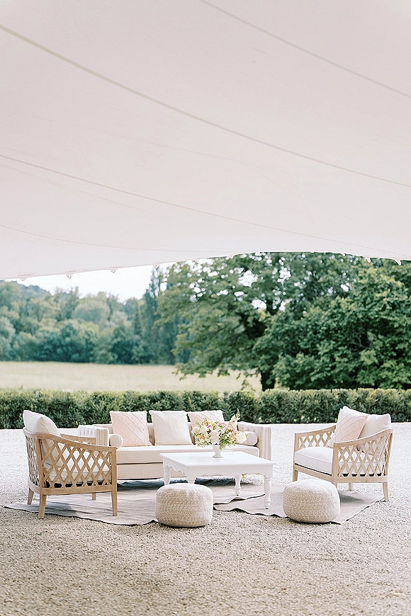 Wedding lounge setup with an outdoor sofa, lounge chairs, and throw pillows around a coffee table under a tent canopy on a gravel patio
