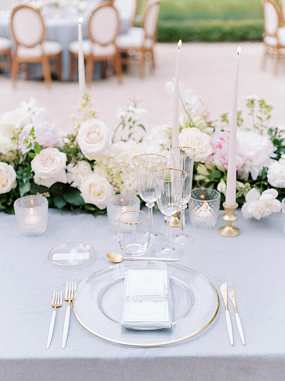 Reception tablescape with wedding place setting, gold rim chargers, brass taper candles, and white rose garland on an outdoor patio table