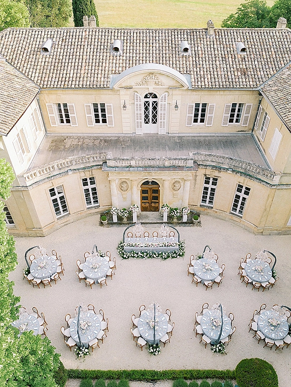 Outdoor reception setup with round tables and a head table draped in floral garland beneath chandeliers in a stone courtyard estate