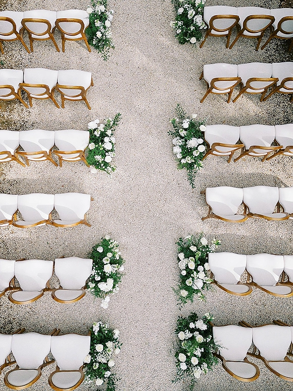 Ceremony aisle decor with symmetrical white flowers and greenery clusters lining a wide gravel aisle beside wooden chairs with cushions
