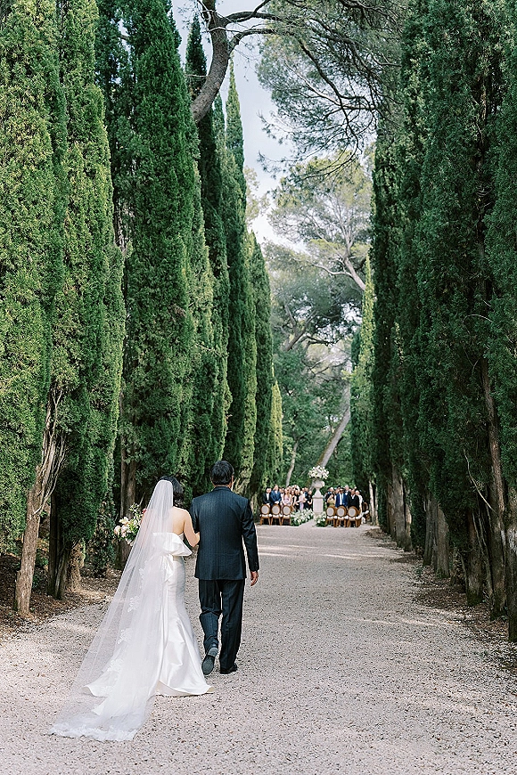 Wedding processional with bride walking down aisle in strapless gown and long veil, holding bouquet as father escorts her along cypress-lined path