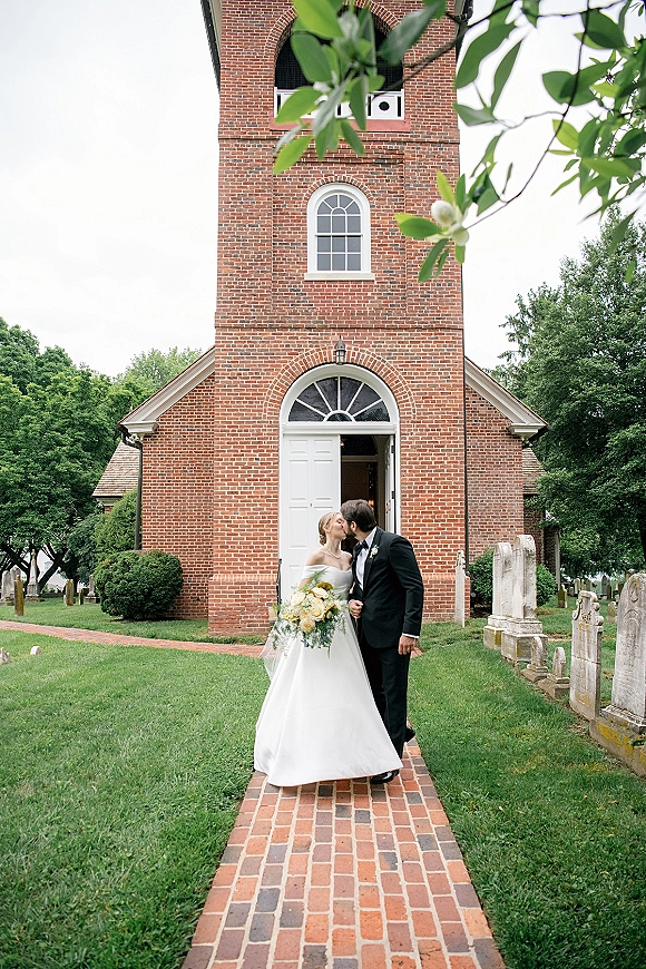 Wedding kiss as bride in strapless dress and groom in black tuxedo embrace with bouquet at brick church doorway near headstones