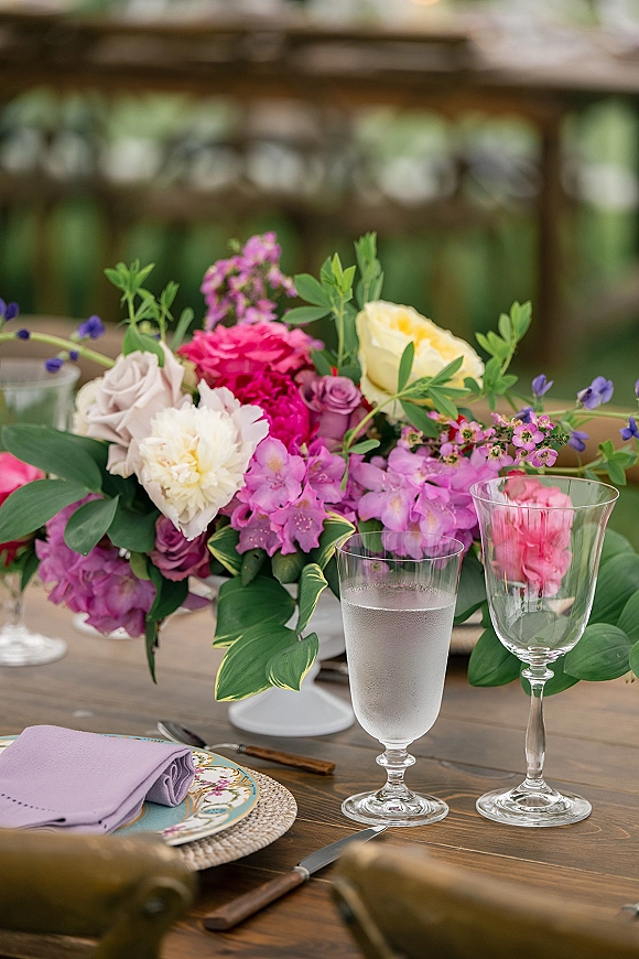 Reception tablescape with colorful wedding centerpiece of roses and hydrangea, clear goblets, patterned plates and lavender napkin on wood table outdoors