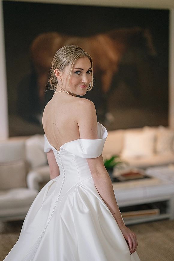 Bridal portrait of a bride in an off the shoulder wedding dress, looking over her shoulder to show button-back satin gown in a living room