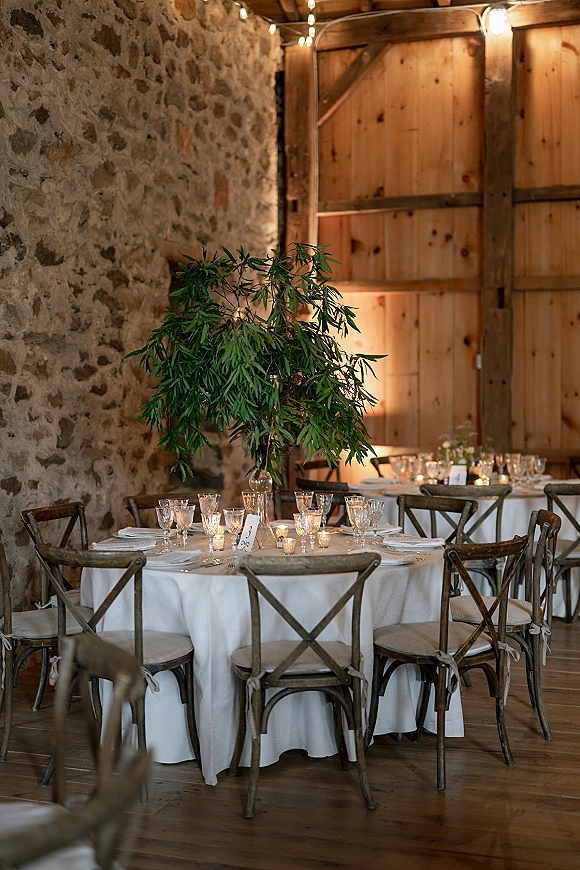 Reception tablescape with a round wedding table setup, white tablecloth, greenery centerpiece, glass goblets, and candlelit string lights by barn doors