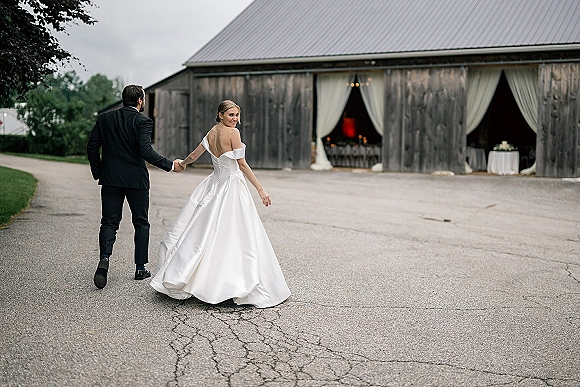 Couple portrait of bride and groom walking away holding hands, her off-the-shoulder gown and veil flowing past a rustic barn entrance