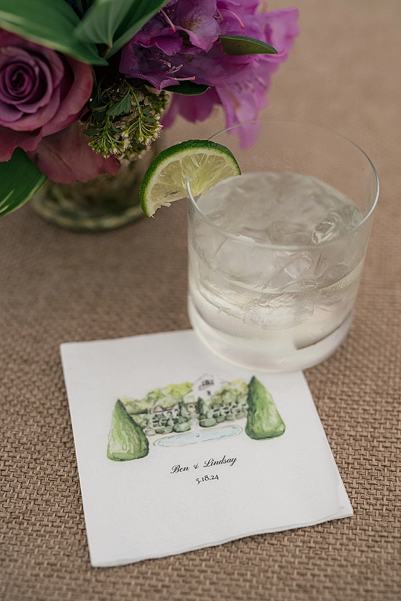 Wedding invitation with watercolor wedding invitation illustration beside a rocks glass cocktail with lime, ice, and purple rose centerpiece on rattan placemat