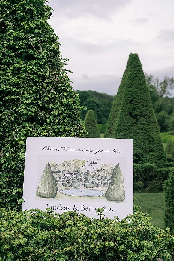 Wedding welcome sign with custom venue illustration and calligraphy on an easel, set before evergreen hedges and topiary trees under a cloudy sky