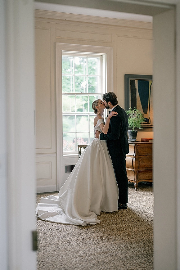 Wedding kiss portrait of bride and groom kissing by a tall window, her off-the-shoulder gown and long veil train flowing across carpeted floor