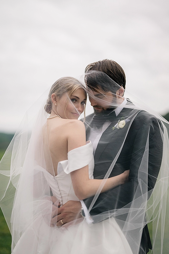 Couple portrait of bride and groom under veil, bride glancing over shoulder in pearl necklace, groom in tuxedo in grassy field under overcast sky