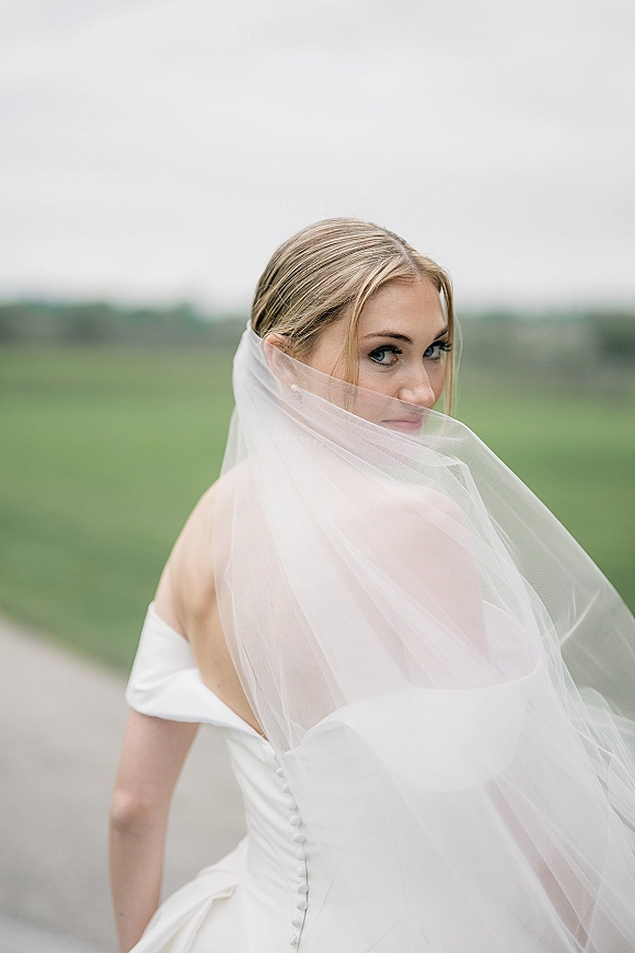 Bridal portrait of a bride looking over shoulder with a long tulle veil over her face on a country road under an overcast sky