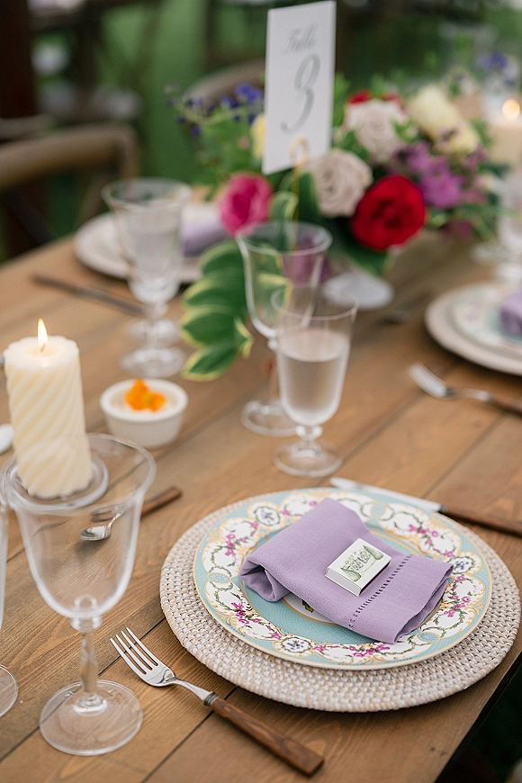 Reception tablescape with wedding table setting featuring colorful floral centerpiece and taper candle on rustic wood table with green chairs behind